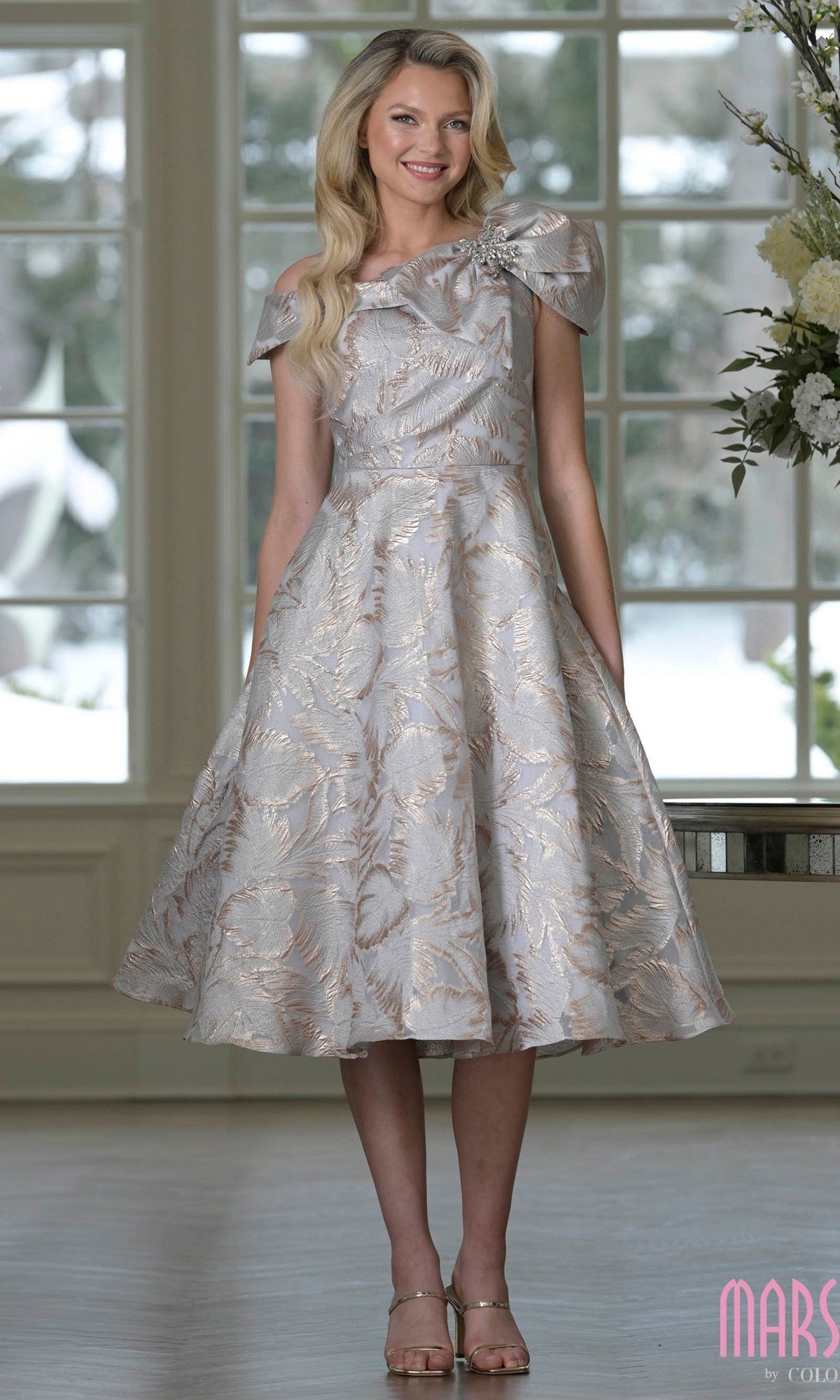 A woman stands indoors wearing the Marsoni MV1413 Knee Length Semi-Formal Dress in gold gray, featuring an off-the-shoulder textured jacquard design with a flared skirt and metallic leaf pattern, paired with high-heeled sandals.